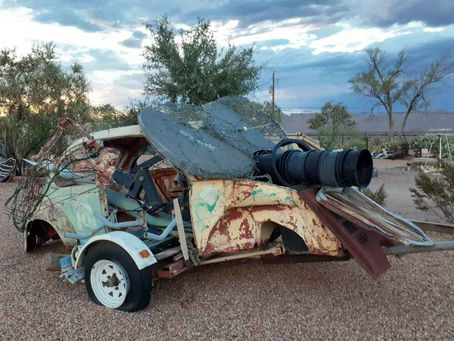 Rusty, dismantled car adorned with a telescope and metal mesh in a desert setting under a cloudy sky. Surrounding trees add an abandoned feel.