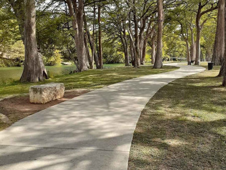 A curved path winds through Louise Hays park with tall trees casting shadows. Sunlit grass and a stone block are visible, creating a serene setting.