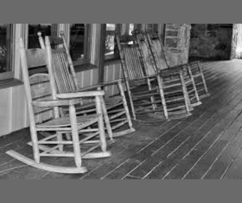 Five empty wooden rocking chairs lined up on a rustic porch with wooden flooring. Stone wall and windows in the background. Black and white.