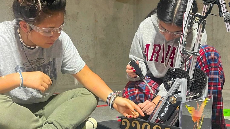 Two people work on a robot with "23669" on it. One wears a gray shirt, the other an "ARVADA" hoodie, both in a concrete room.