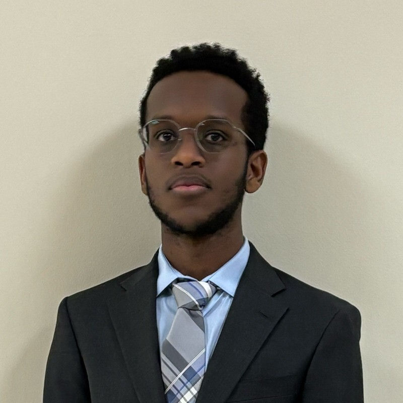 Young man in glasses and suit with a plaid tie stands against a plain wall, looking confident and composed.