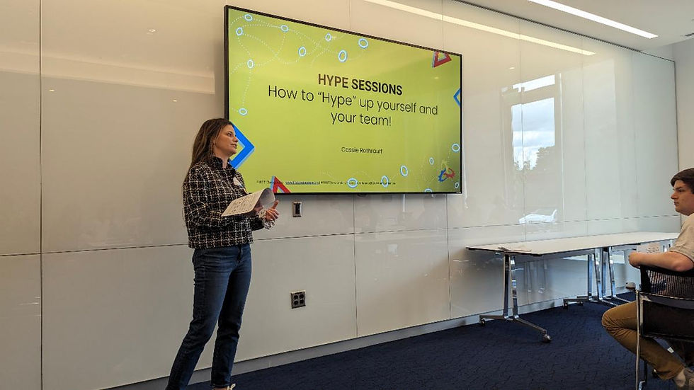 A woman presents in a modern room, gesturing with notes. Screen reads "Hype Sessions: How to 'Hype' up yourself and your team!" Audience listens.
