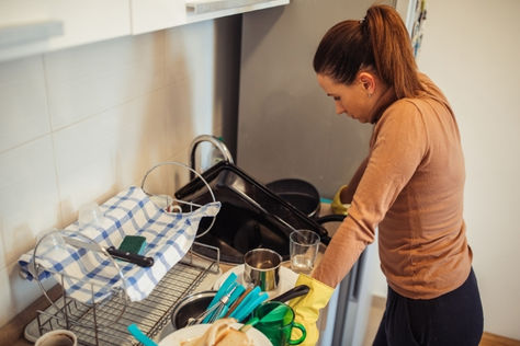 A woman takes a contemplative pause while standing at a kitchen sink full of dirty dishes. She is wearing yellow rubber gloves.