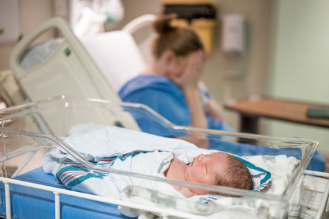 A newborn baby sleeps in a hospital bassinet, with a mother in the background covering her face in distress, depicting perinatal trauma, postpartum mental health struggles, or emotional recovery after childbirth.