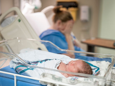 A newborn baby sleeps in a hospital bassinet, with a mother in the background covering her face in distress, depicting perinatal trauma, postpartum mental health struggles, or emotional recovery after childbirth.