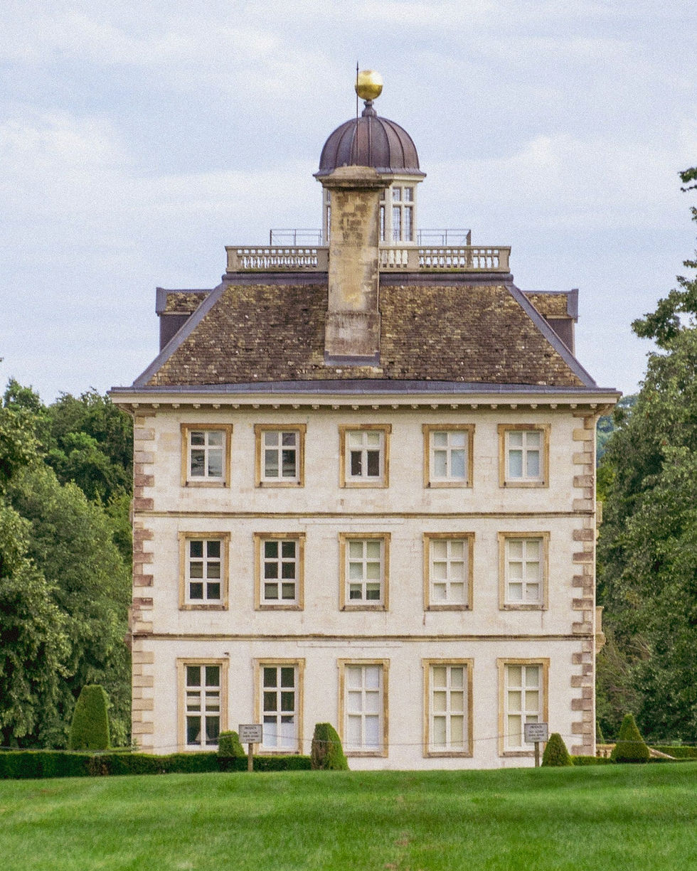 A classic three-story stone building with a domed chimney, surrounded by lush greenery. The sky is overcast, and the grass is vibrant green.