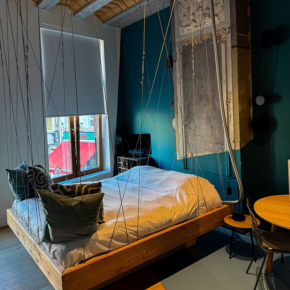 Suspended wooden bed with white bedding in a modern room with teal walls. A large window with blinds lets in soft light. Cozy atmosphere.