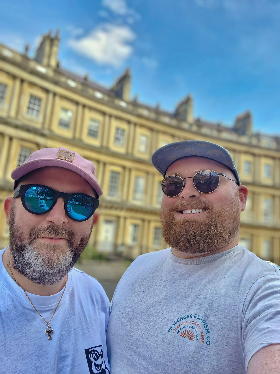 Two men with sunglasses smile for a selfie in front of a historic yellow building under a blue sky. One wears a cap and the other a cross necklace.