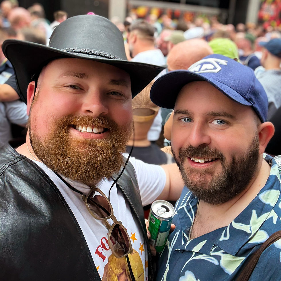 Two smiling men at a crowded outdoor event, one in a cowboy hat and T-shirt, the other in a cap and patterned shirt, holding a drink can. Pride Event London