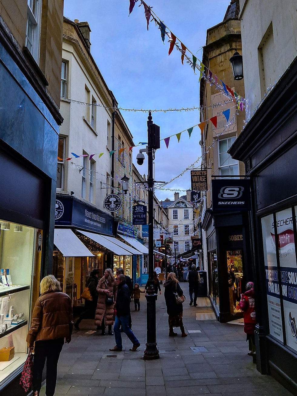 Narrow street with shops, people walking and browsing, colorful bunting overhead, evening setting, signs for Neal's Yard and Sobeys visible.