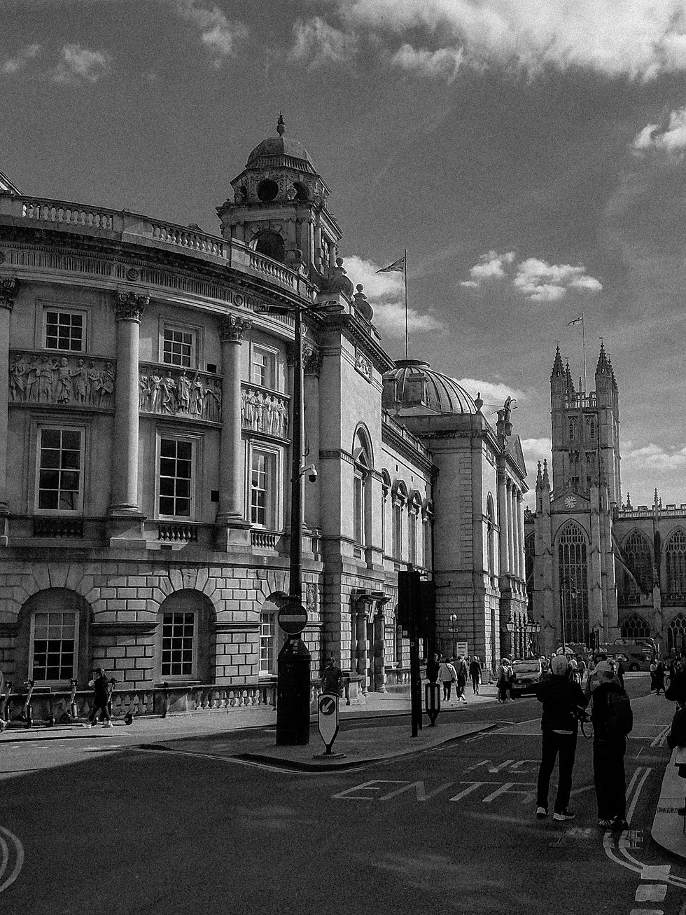 Street scene with people walking, surrounded by historic buildings featuring columns and sculptures. Cloudy sky sets a timeless mood.