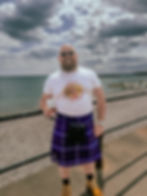 Man in sunglasses and kilt smiles at seaside holding a drink. Shirt reads "Dolly for President." Cloudy sky, relaxed mood.