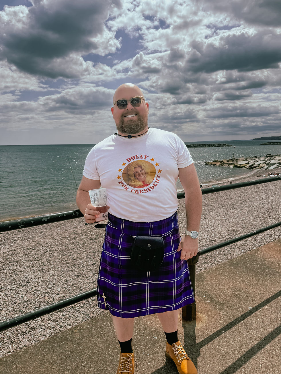 Man in sunglasses and kilt smiles at seaside holding a drink. Shirt reads "Dolly for President." Cloudy sky, relaxed mood.