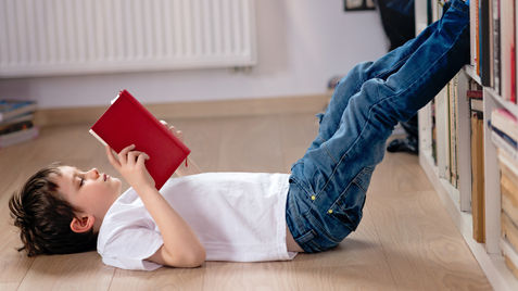 A young boy lays on his back reading a red book.