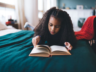 Elementary school girl reading book while lying on bed