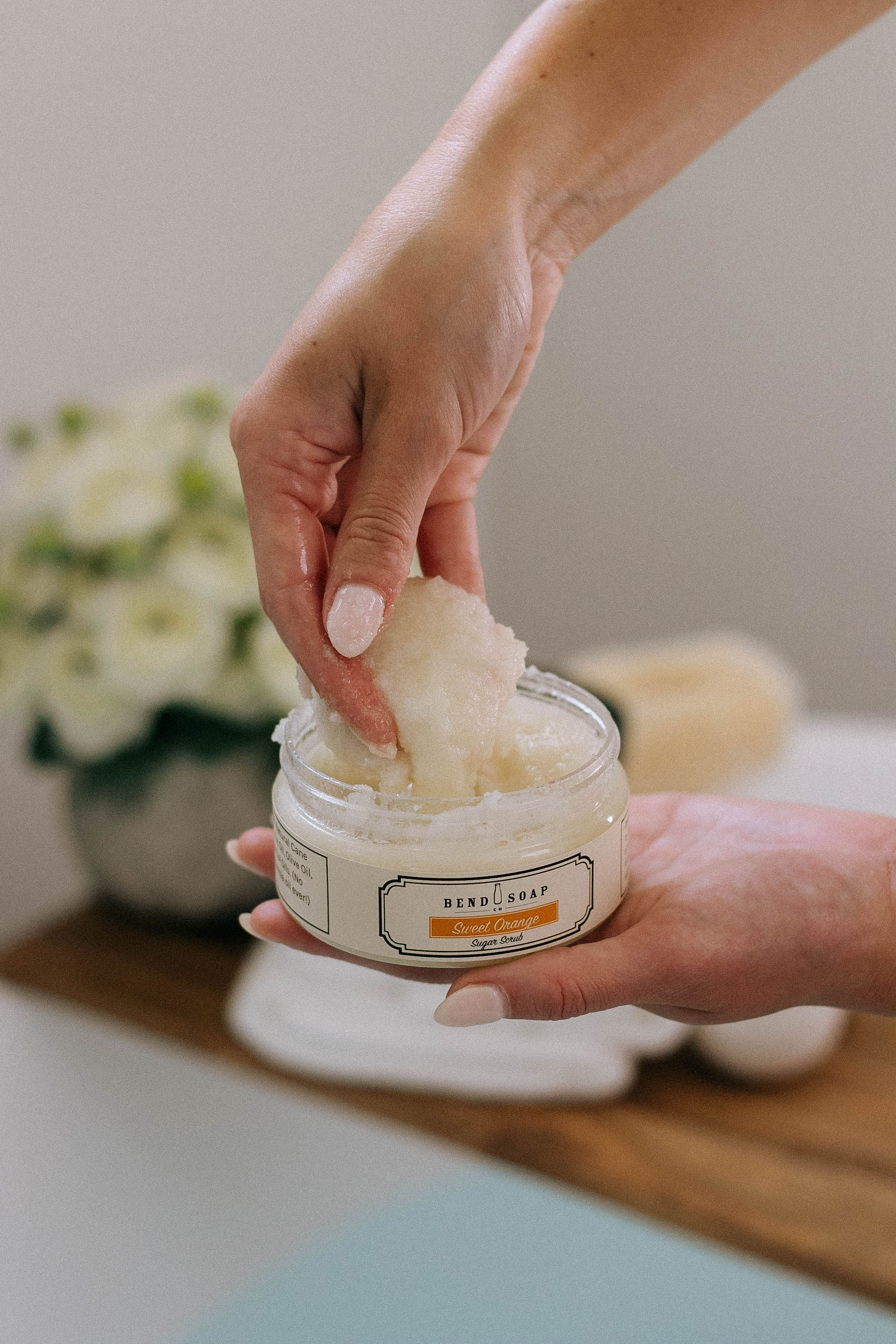 Hand scooping Bend Soap Co. Sweet Orange Scrub out of jar with white potted flowers and wooden bathtub plank in backdrop