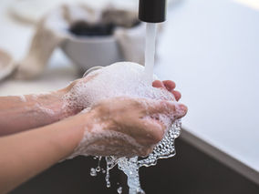 Two hands full of soap suds under running water in farmhouse sink.