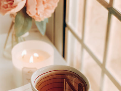 A cup of tea, a burning candle, and a vase of peonies at a window's ledge.