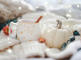 Teacup with cinnamon stick next to white pumpkin surrounded by fairy lights, fall flowers, and leaves.