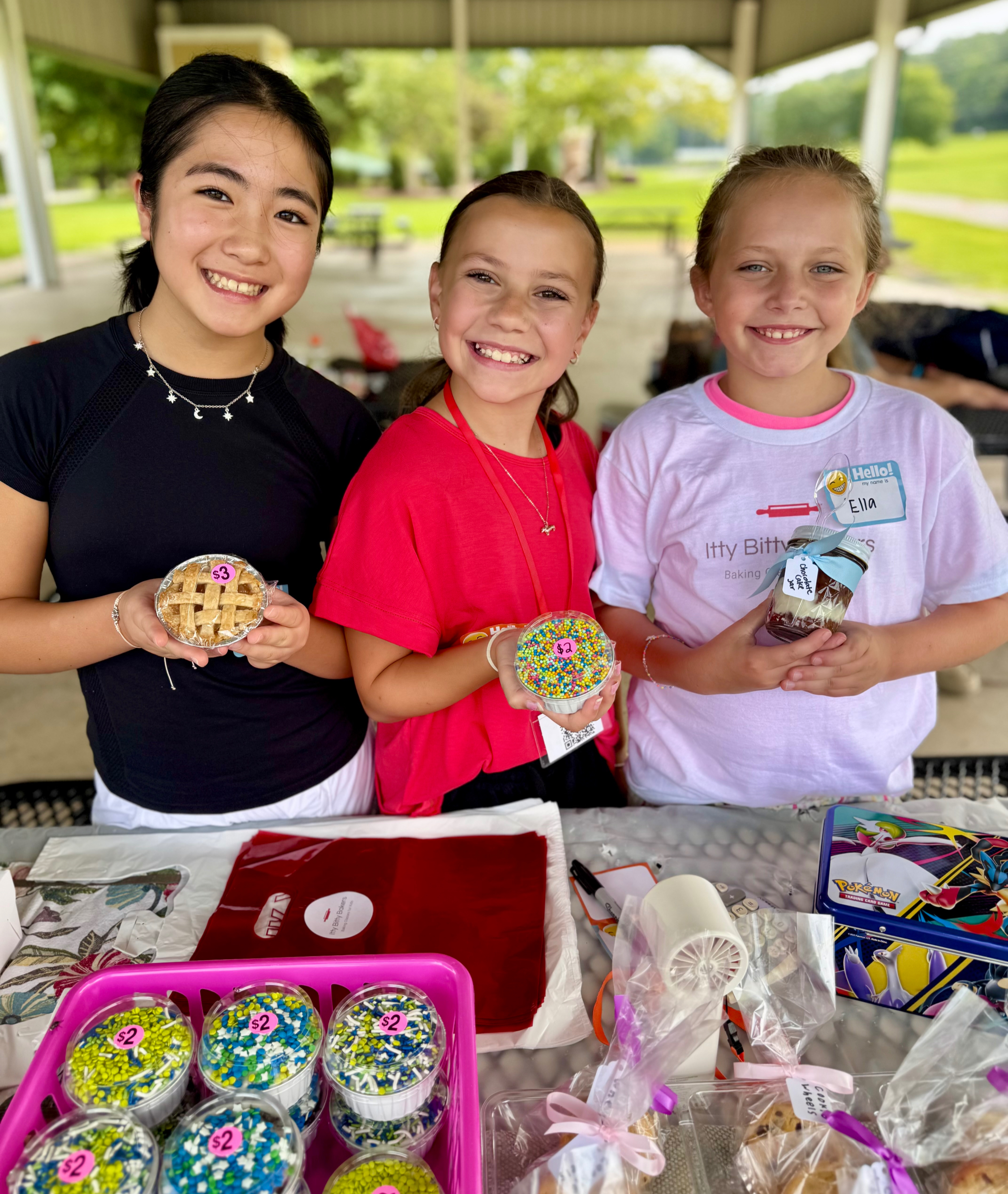 Three smiling girls at a park pavilion bake sale hold pies, sprinkles, and a jarred dessert with price tags and name stickers.