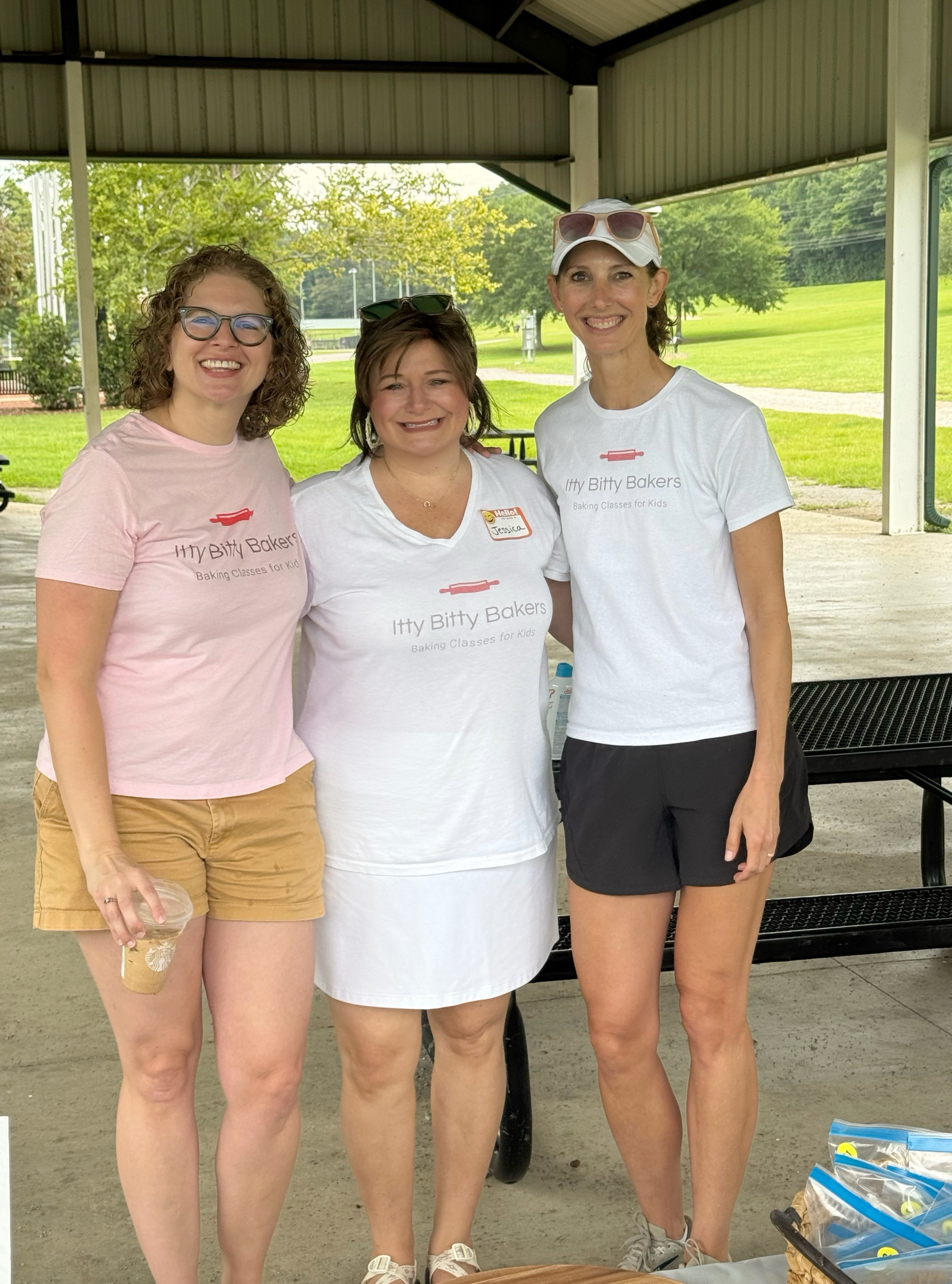 Three smiling women in Itty Bitty Bakers shirts stand under a park pavilion, with green grass and trees behind them.