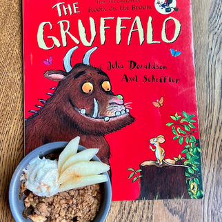 The Gruffalo book with red cover on a wooden table, next to a bowl of crumble topped with cream and sliced apples.