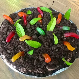 Dirt cake with crumbled chocolate, colorful gummy worms, and green mint leaves on top, set on a wooden table.