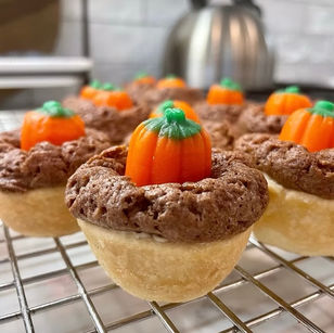 Mini pumpkin cupcakes with brown frosting and candy pumpkins on a cooling rack. Background shows a silver kettle and white tiles.