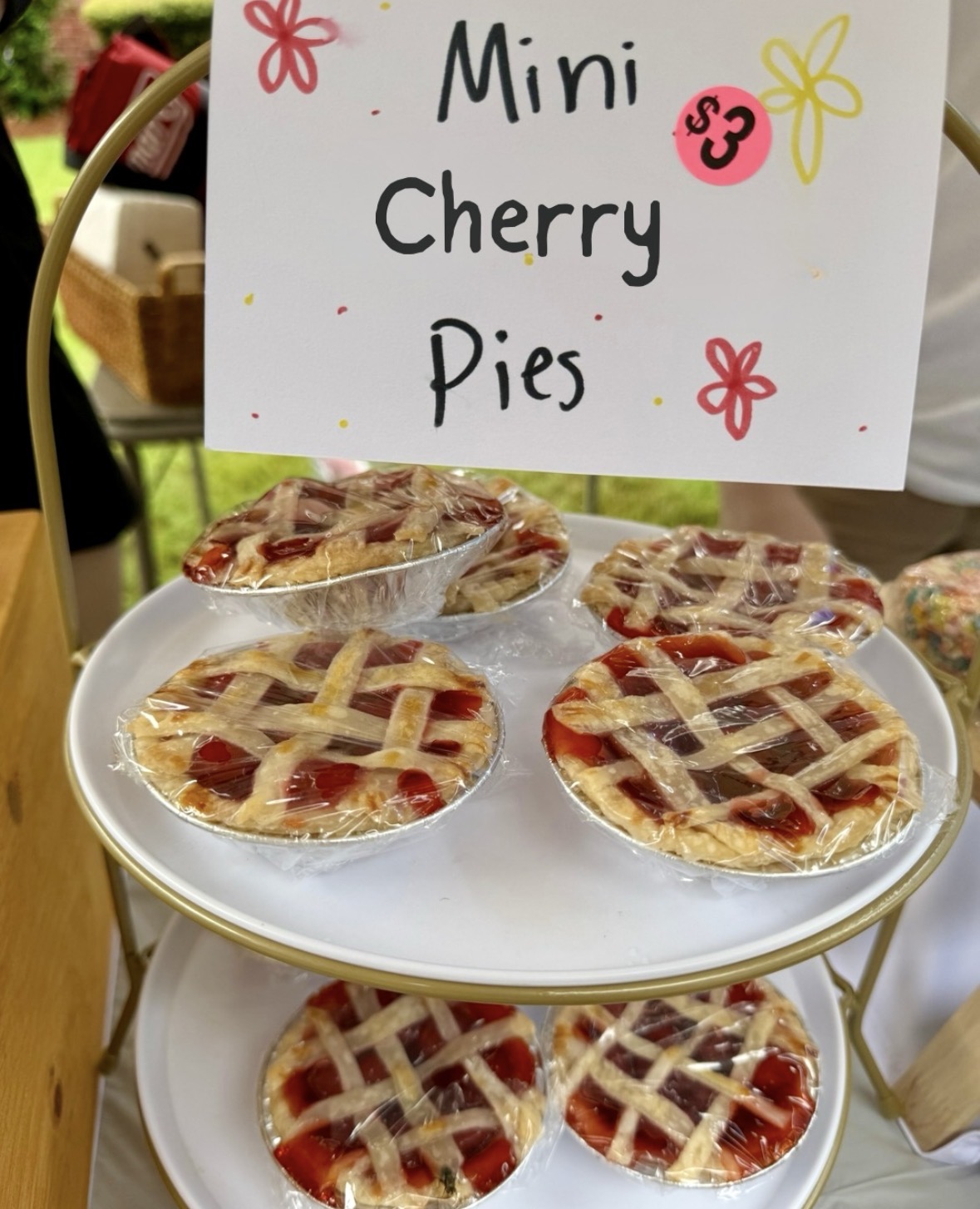 Mini cherry pies on a white tray cart with a pink $3 sign, wrapped in plastic at an outdoor market.