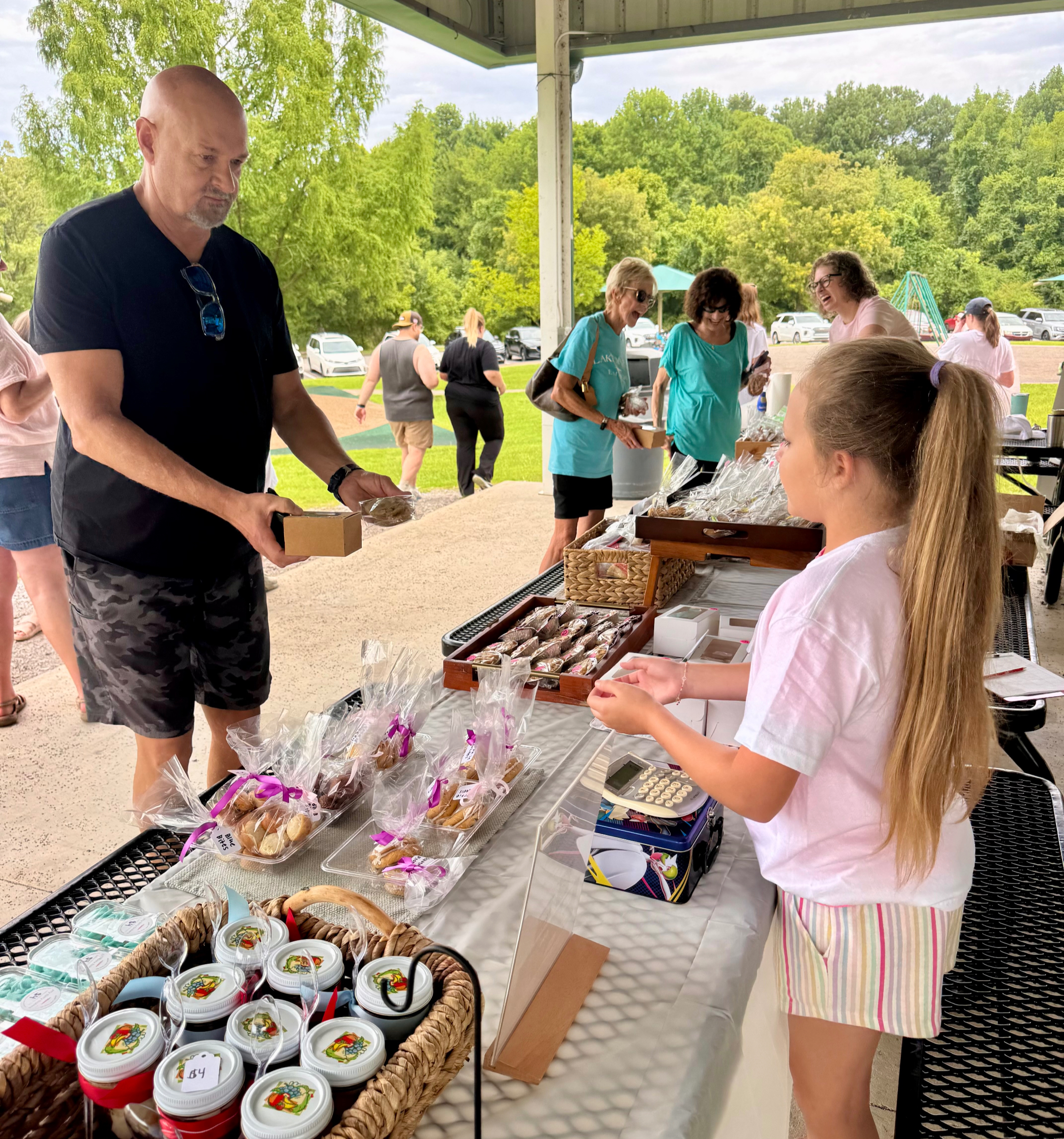 Outdoor bake sale under a pavilion, with a girl helping a man buy packaged treats while shoppers mingle and trees fill the background.