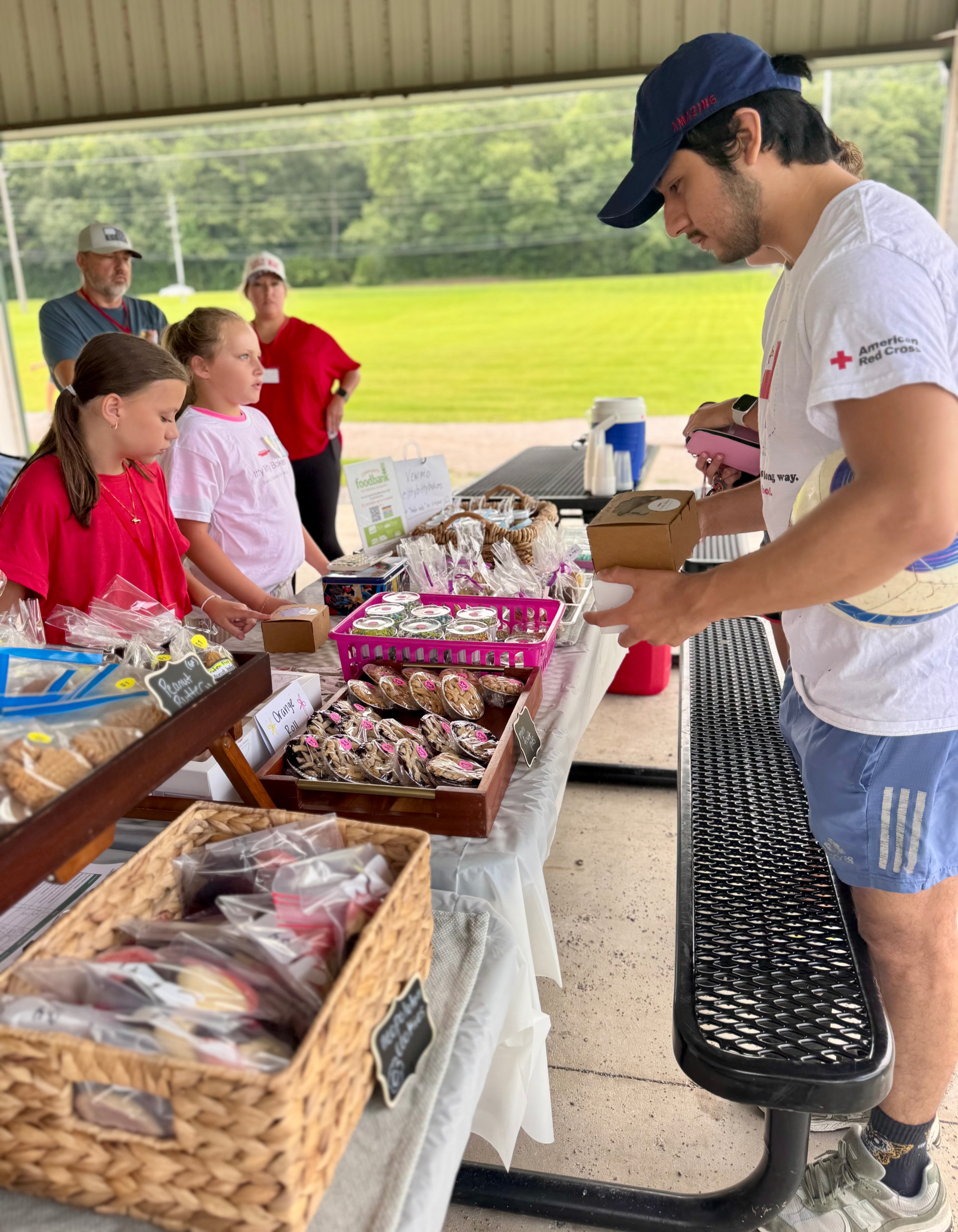 People browse baked goods at a picnic table stall; a man in an American Red Cross shirt pays, outdoors under a pavilion.