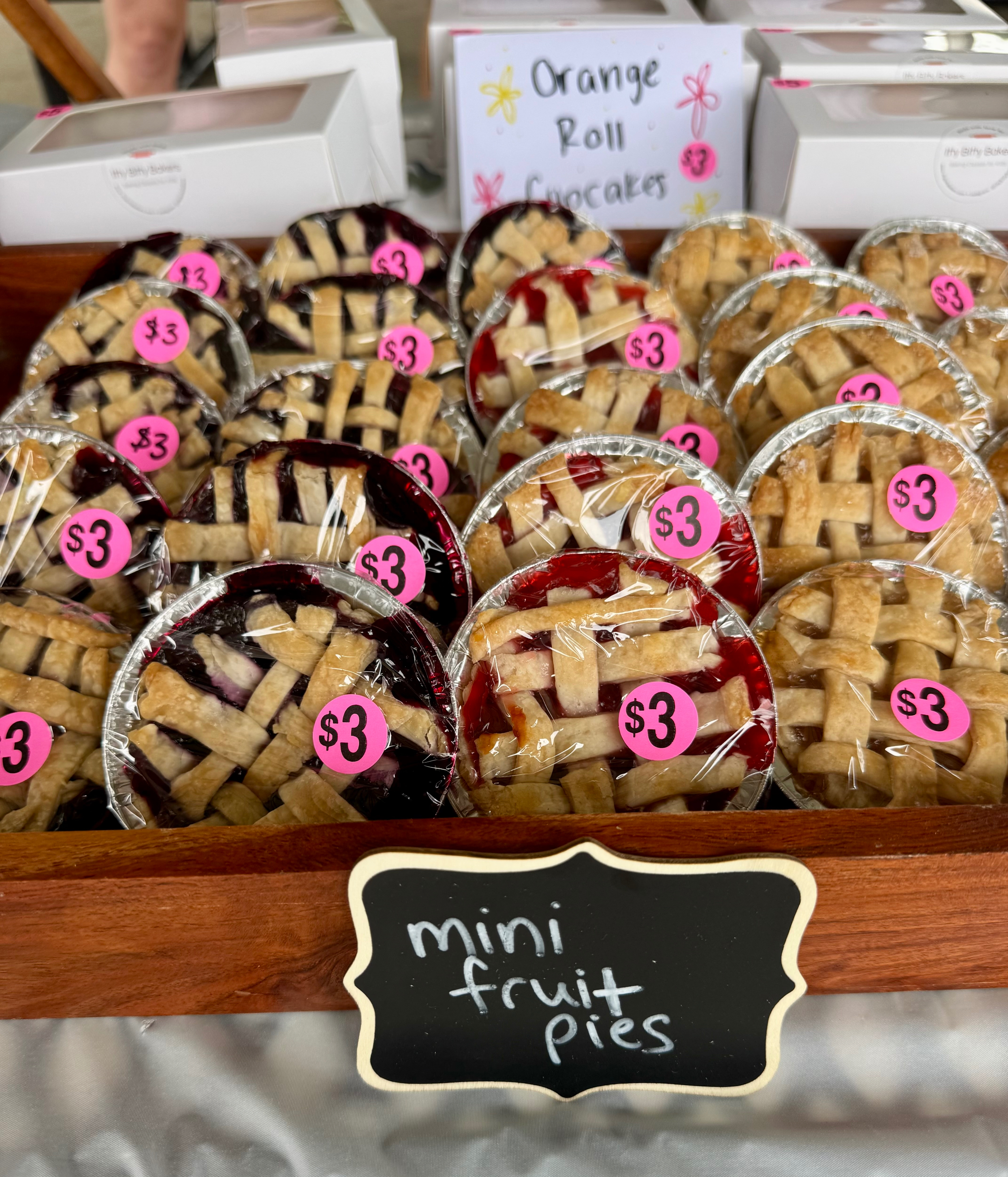 Tray of mini fruit pies with lattice crusts and $3 pink stickers; Orange Roll Cupcakes sign in the background.