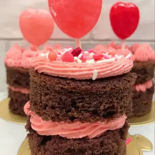 Chocolate cake with pink frosting and sprinkles, topped with red heart-shaped toppers, set against a light brick background.