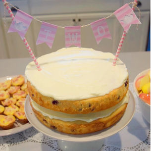 Two-layer cake with white frosting on a stand, pink banner reading "TWO," surrounded by snacks on a lace-patterned tablecloth.