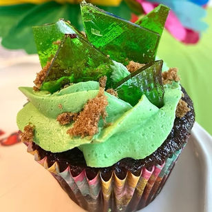 Chocolate cupcake with green frosting, green sugar shards, and brown crumbs. Background has bright, colorful paper decor.