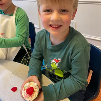 A smiling child in a green Grinch shirt holds a cupcake with white frosting and red sprinkles. Another child in an apron is nearby.