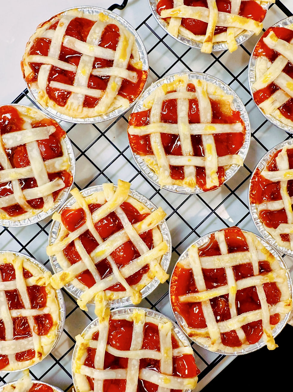 Mini cherry lattice pies in foil tins cooling on a black rack, with glossy red filling and golden crusts.