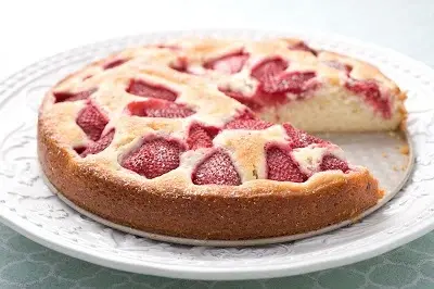 Strawberry cake with a slice cut out, on a decorative white plate. Bright red strawberries contrast with the light, airy cake.