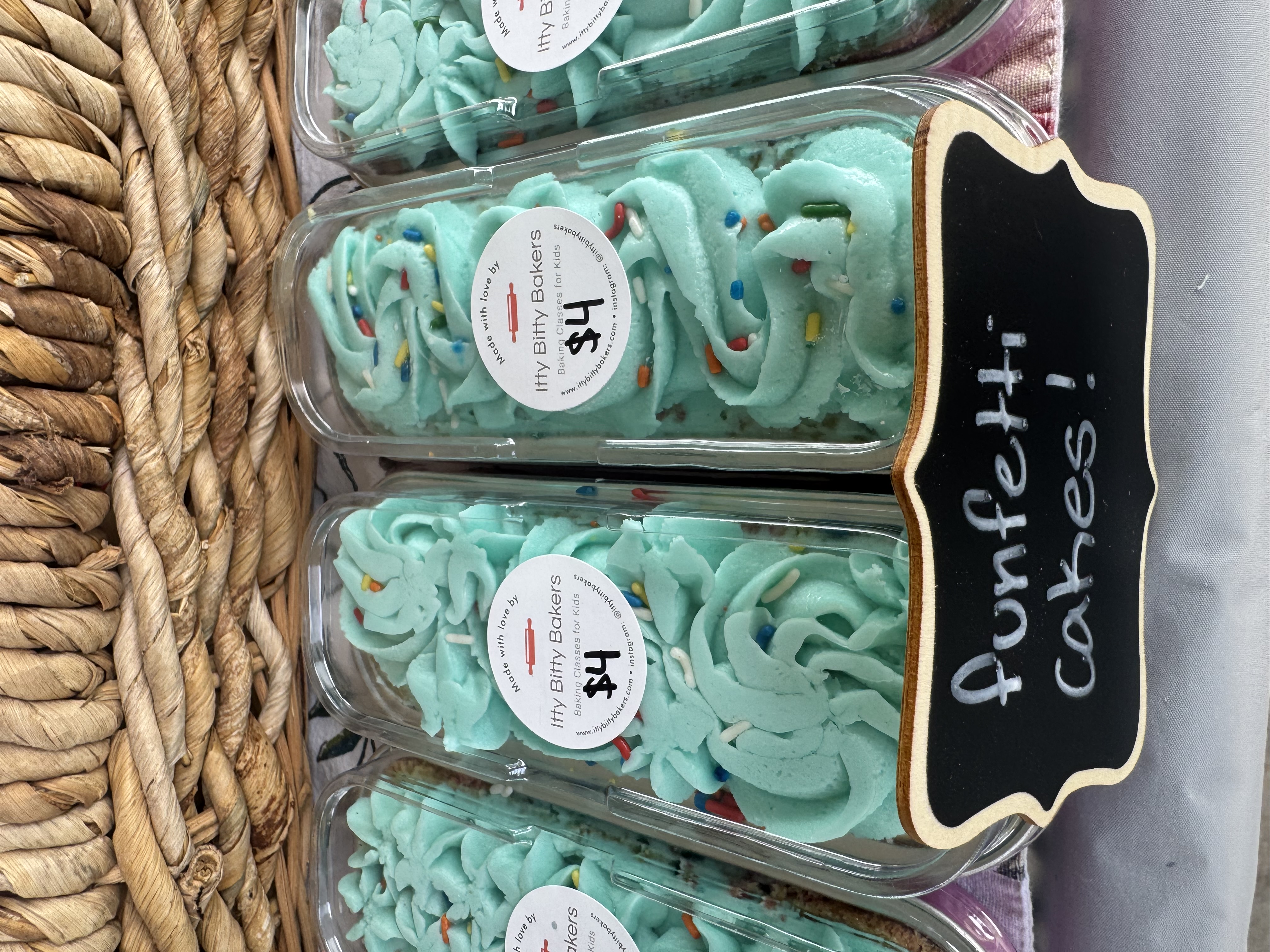 Turquoise funfetti cakes in clear trays labeled Itty Bitty Bakers $4, with a woven basket backdrop