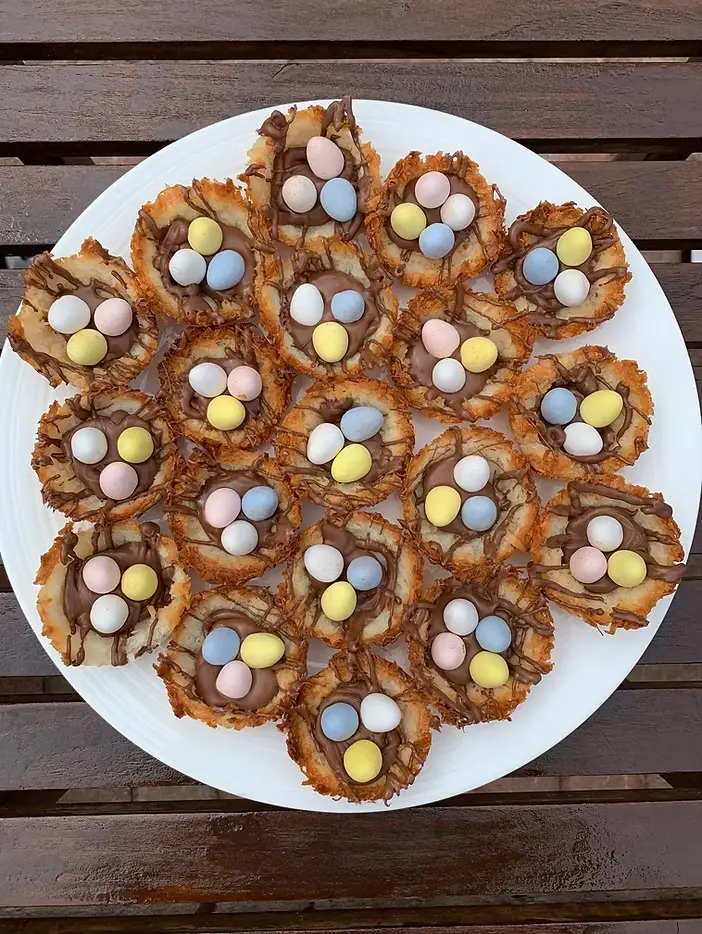 Chocolate nests with pastel candy eggs on a white plate, set on a wooden table. The colorful treats evoke a festive mood.