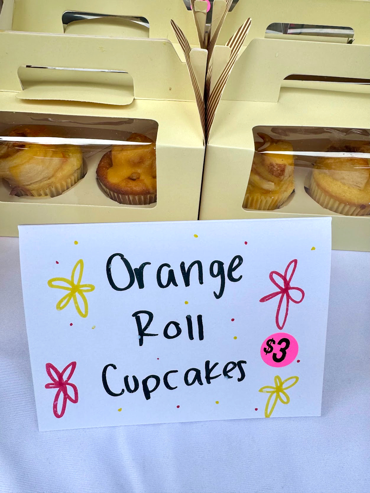 Cream-colored bakery boxes of orange roll cupcakes behind a handwritten sign reading Orange Roll Cupcakes, $3