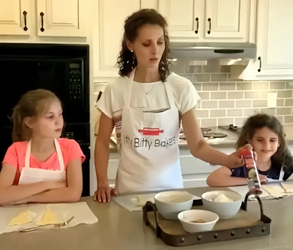 Woman and two girls baking in a kitchen. Kitchen features white cabinets and tiled backsplash.