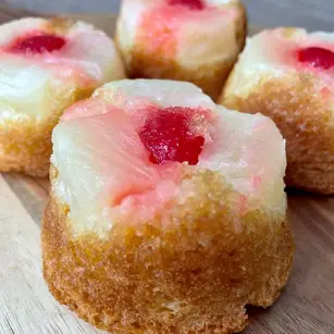 Four mini pineapple upside-down cakes on a wooden surface, topped with red cherries. Warm tones, moist texture, sweet and inviting.