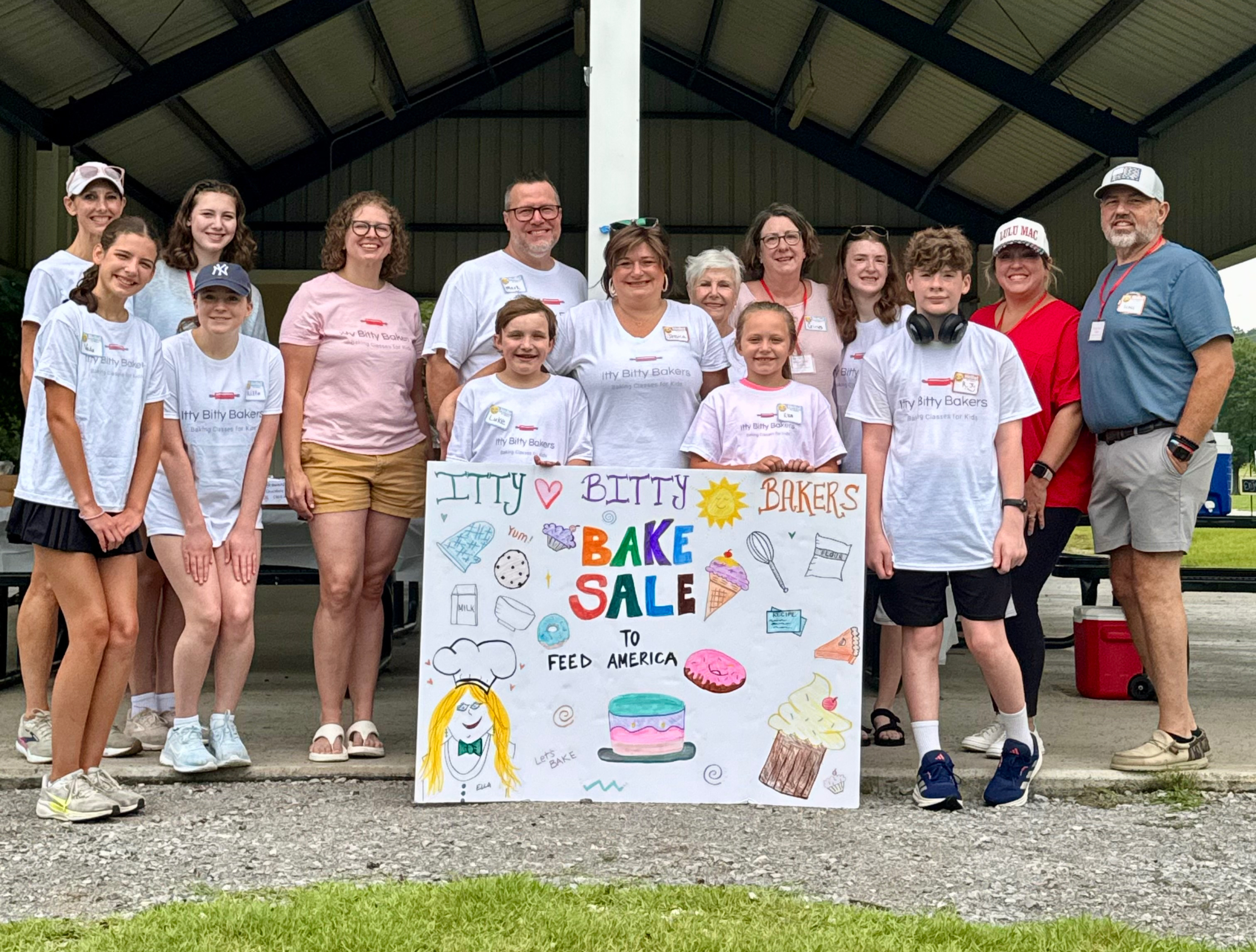Group of kids and adults posing under a pavilion with a colorful sign reading Itty Bitty Bakers Bake Sale to Feed America.