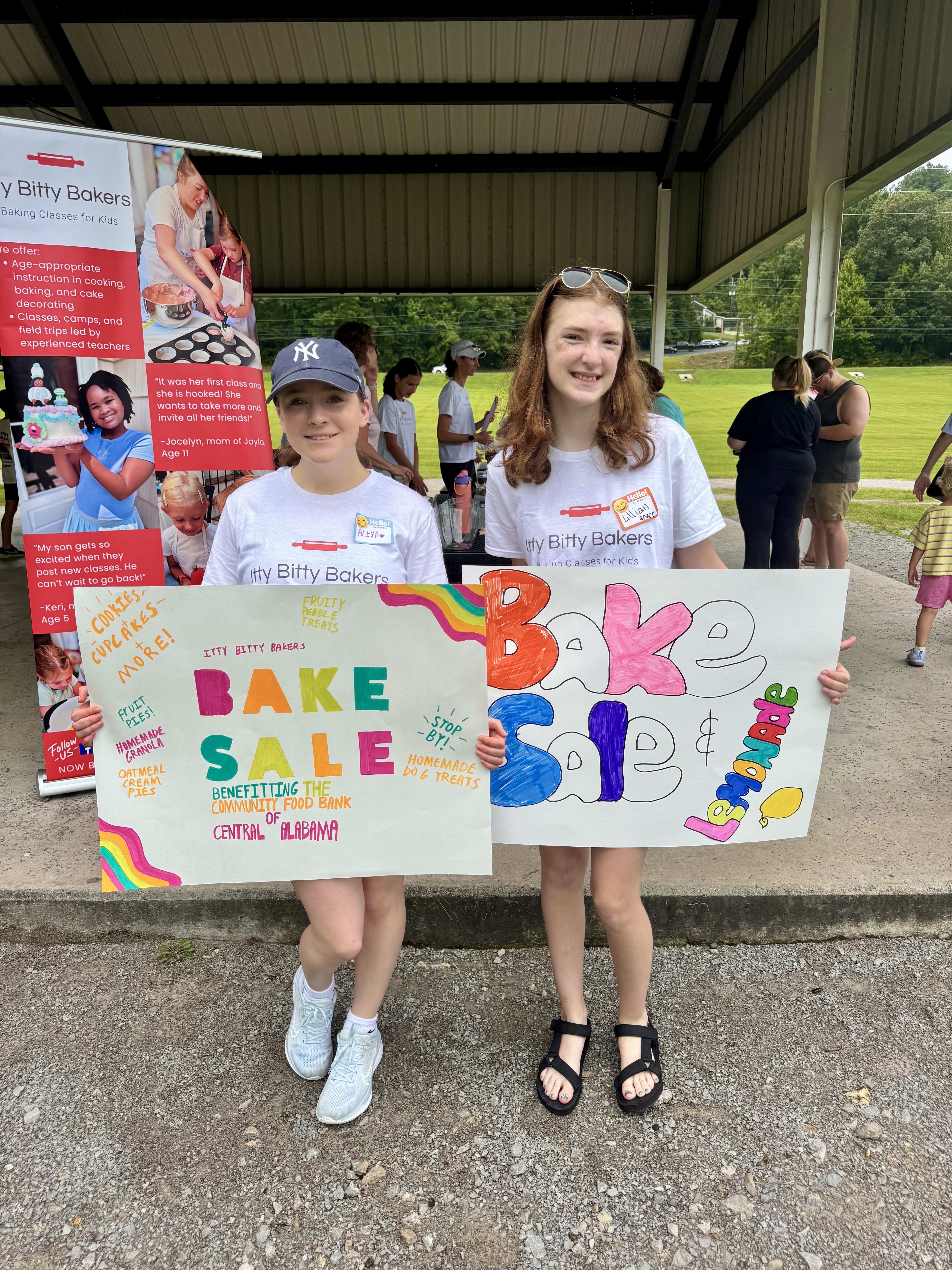 Smiling girls hold colorful Bake Sale signs for Itty Bitty Bakers at a pavilion, with a promotional poster behind them.