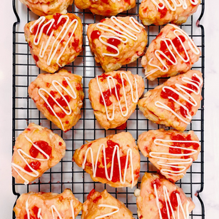 Heart-shaped scones topped with red jam and white icing on a cooling rack, set against a white marble background.
