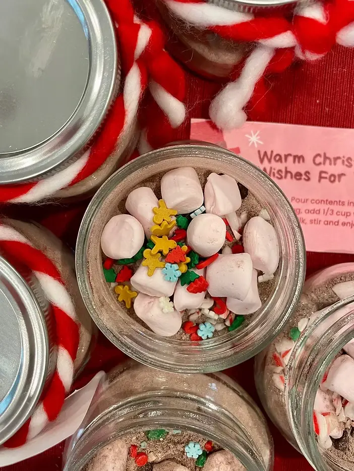Jars filled with cocoa mix, pink marshmallows, and colorful sprinkles. Red and white yarn lids. Pink card reads "Warm Christ...".