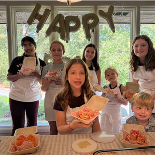 Kids in aprons hold decorated cookies in boxes, smiling indoors. Balloons spell "HAPPY" above, and a green backdrop is visible outside.