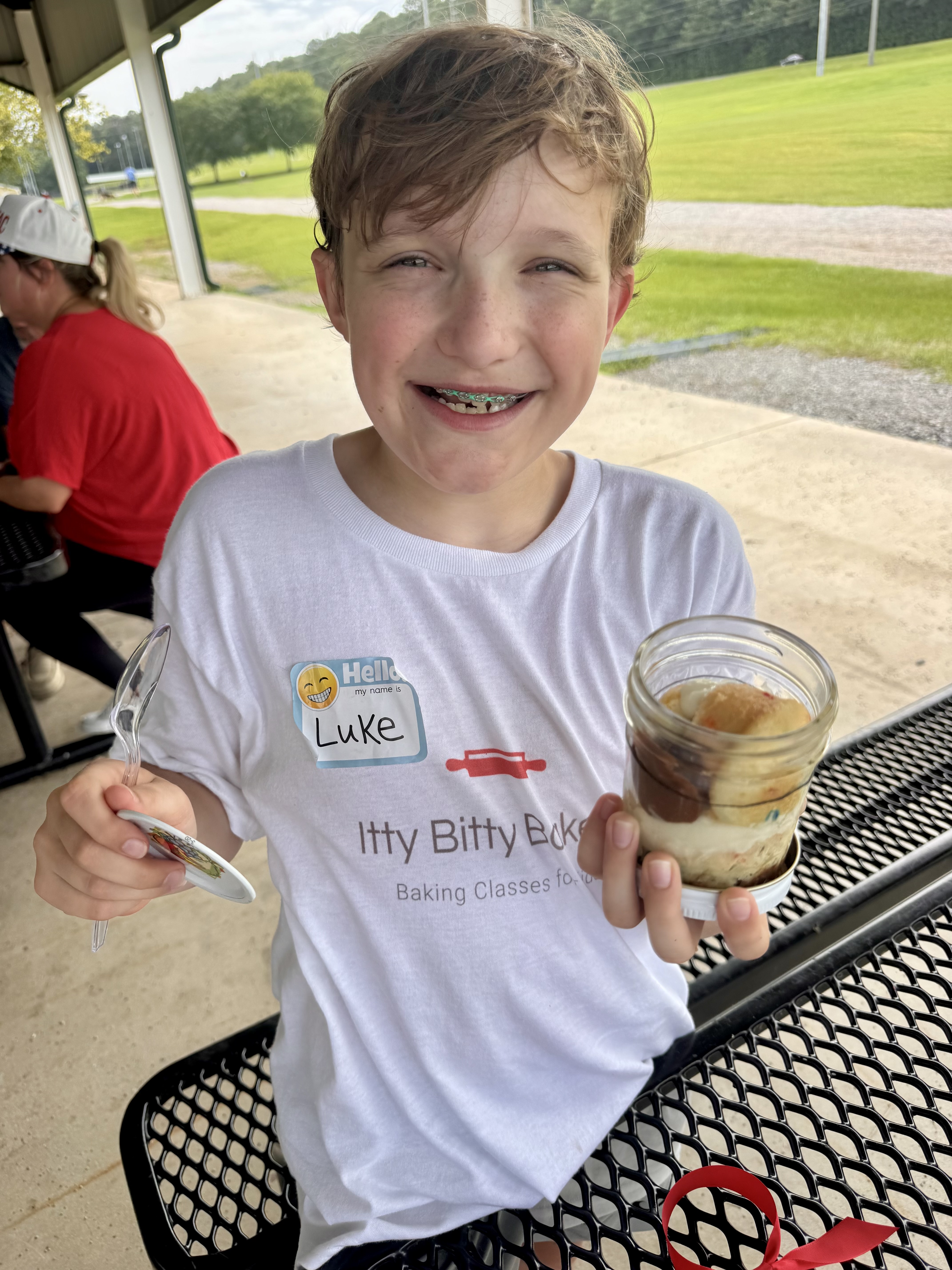Smiling boy with braces, name tag Luke, holds a dessert cup and spoon at a picnic table in a sunny park.