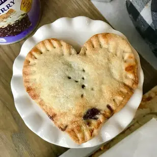 Heart-shaped pastry on a white plate, wooden table background. Nearby, a can reads "berry." Cozy kitchen vibe with a checkered cloth.